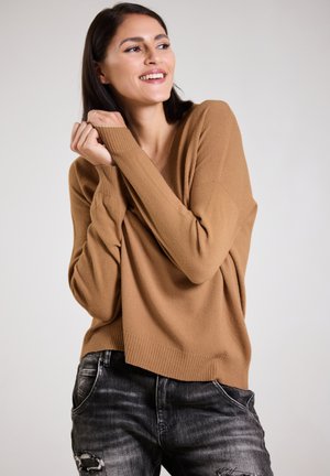 Kamel-farbener, oversized V-Ausschnitt-Pullover aus weichem, strukturiertem Stoff, kombiniert mit dunkelgrauen, lässigen Jeans mit Rissen. Das Design zeichnet sich durch lange Ärmel und einen lässigen Schnitt aus.