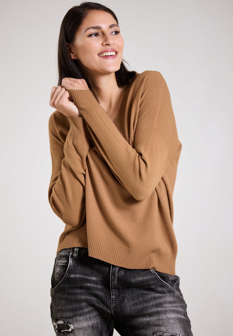 Camel-colored, oversized v-neck sweater in soft, textured fabric, paired with dark gray ripped jeans. The design features long sleeves and a relaxed fit.