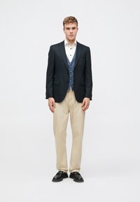 Dark blazer over a light blue vest and white shirt, paired with beige trousers and black shoes. Smooth fabric, tailored fit.