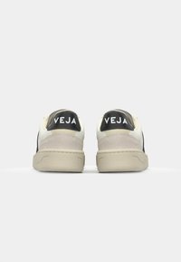 White and black sneakers featuring a textured suede heel, flat beige sole, and prominent "VEJA" logo on the back.