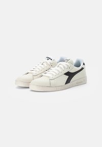 White leather sneakers with black accent, round toe, perforated design, flat rubber sole, and lace-up closure. Lightly textured finish.