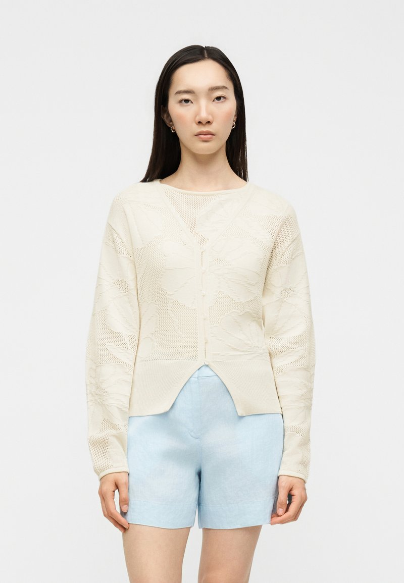 Femme aux longs cheveux foncés porte un cardigan crème en tricot boutonné avec des motifs floraux et un short taille haute bleu clair, debout sur un fond blanc.