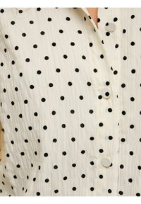 Blouse blanche texturée avec petits pois noirs et boutons recouverts de tissu sur le devant.