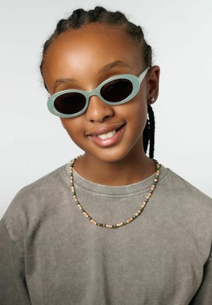 Lunettes de soleil ovales vertes avec des verres foncés, portées avec une chemise grise. Un collier en perles colorées orne le cou, présentant plusieurs nuances.