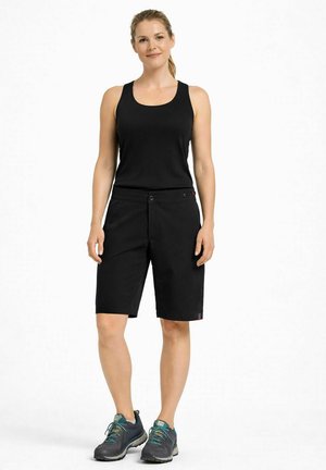 Femme debout, portant un haut noir sans manches, un short noir arrivant aux genoux et des chaussures de randonnée foncées, sur un fond blanc uni.