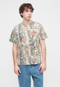 Μπλουζάκι με καμουφλάζ σε μπεζ χρώμα με πράσινα και καφέ μοτίβα φύλλων. Διαθέτει στρογγυλή λαιμόκοψη και πορτοκαλί λογότυπο "Obey" στο στήθος.