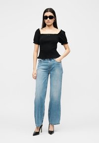 Kvinne iført svart topp med firkantet hals og pufferm, lyseblå høytlivsjegns, svarte hæler og solbriller mot ensfarget bakgrunn.