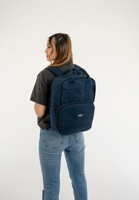 Zaino blu navy con manici doppi, spallacci imbottiti e una tasca frontale con zip. Realizzato in tessuto resistente con una texture liscia.