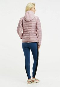 Lichtroze pufferjack met horizontale stiksels en een capuchon, gedragen met blauwe leggings met zwarte zijpanelen en lichtgekleurde sandalen.
