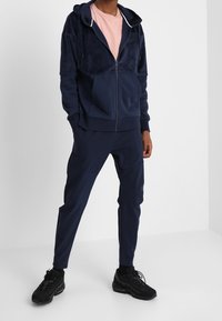 Marinblå fleece hoodie med dragkedja fram, elastiska ärmslut och rosa T-shirt under, i kombination med matchande marinblå joggers och svarta sneakers.