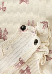 Vaate vaaleasta puuvillasta, jossa on ribbauskudonta ja vaaleanpunaisia nauhakuviota. Sisältää mustat nepparit ja röyhelöreunan koristeellinen yksityiskohtana.