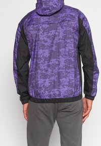 Lila kamouflagejacka med svarta ärmar, elastisk i midjan och vid ärmsluten, samt med huva. Tillverkad av lätt, strukturerad tyg.