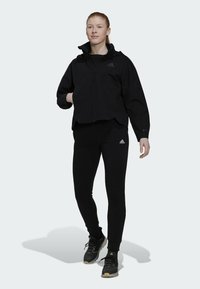 Zwarte windbreaker met een hoge kraag, gecombineerd met zwarte leggings en sneakers. Heeft een gemêleerde stof en een Adidas-logo op de borst.
