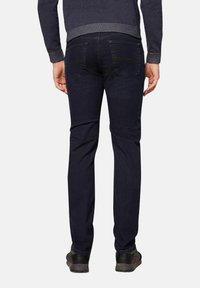 Donkerblauwe jeans met een slim fit, vijf-pocketontwerp en subtiele stikdetails, gecombineerd met zwarte schoenen en een grijs shirt.