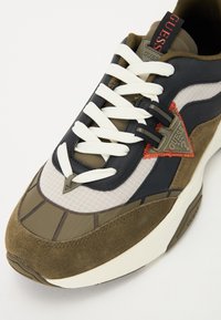 Sneaker, amely különböző anyagok keverékéből készült, beleértve a sötétkék bőrt, olíva színű velúrt és hálós szövetet. Fehér fűzőkkel, piros kiegészítőkkel és háromszög alakú logóval rendelkezik.