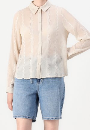 Osoba ubrana w beżową bluzkę zapinaną na guziki z koronkowymi detalami oraz jasnoniebieskie dżinsowe szorty, stojąca z jedną ręką na biodrze na jednolitym tle.