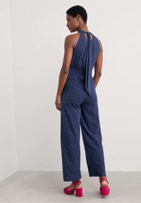 Tumed jumpsuit valgete ringidega, kaelus on halter-stiilis ja seljal on sidumisdetail. Lahtine lõige ja laiad sääred. Kombineeritud erkkollaste plokk-kontsadega.