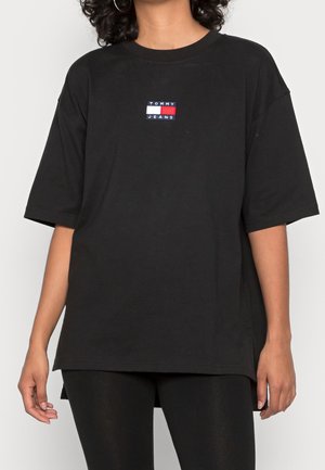 Femme portant un t-shirt noir surdimensionné Tommy Jeans avec un patch logo centré sur la poitrine, associé à un legging noir.