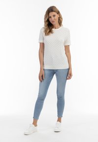 Hvid, tekstureret sweatshirt med korte ærmer og diamantmønster, parret med lyseblå skinny jeans og hvide sneakers. Ensfarvet baggrund.