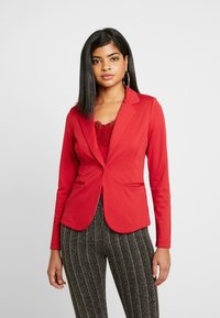 Blazer vermelho com um design ajustado, fecho de botão único e dois bolsos frontais. Usado sobre uma blusa de renda, combinado com calças pretas com padrão.