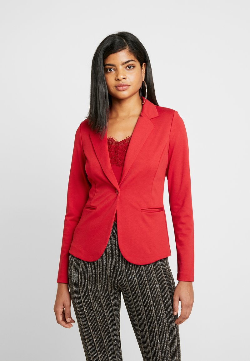 Blazer vermelho com um design ajustado, fecho de botão único e dois bolsos frontais. Usado sobre uma blusa de renda, combinado com calças pretas com padrão.