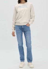 Sudadera beige clara con "OPEN HEARTS OPEN MINDS" bordado en azul, combinada con vaqueros azul claro y zapatillas blancas. Corte estándar.