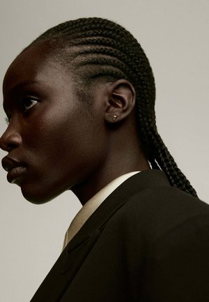 Vista de perfil de una persona con el cabello trenzado, que lleva una chaqueta de traje oscura sobre una camisa de color claro. Pendientes en forma de estrella pequeños son visibles.