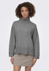 Suéter gris de gran tamaño con un cuello alto en relieve y dobladillo acanalado. Textura suave, mangas largas, llevado sobre una falda estampada.