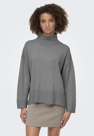 Suéter gris de gran tamaño con un cuello alto en relieve y dobladillo acanalado. Textura suave, mangas largas, llevado sobre una falda estampada.