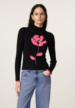 Vrouw die een zwarte trui met lange mouwen draagt met een groot roze bloemenpatroon en blauwe spijkerbroek, staand met één hand in haar zak.