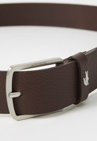 Brązowy skórzany pasek o teksturowanym wykończeniu, srebrna klamra metalowa z wygrawerowanym napisem "LACOSTE", z małym emblematem krokodyla na pasku.