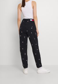 Vaqueros de mezclilla negra con un patrón de corazones bordado, corte ajustado y cintura alta. Combinados con zapatillas blancas y una camiseta sin mangas.