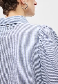 Ριγέ μπλούζα με μπλε και λευκές κάθετες ρίγες, διαθέτει γιακά και φουσκωτά μανίκια. Μικρό λογότυπο στο πίσω μέρος του γιακά.