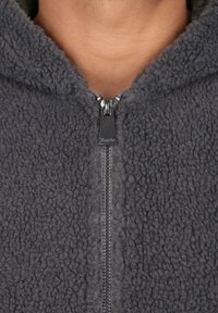 Primo piano di una giacca in pile grigio scuro con la cerniera parzialmente chiusa, che mostra il tessuto testurizzato e parte del collo di una persona.