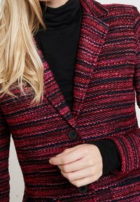 Blazer testurizzato in un vivace rosa e strisce nere, con colletto a punta e chiusura con un solo bottone. Indossato sopra una maglia nera.