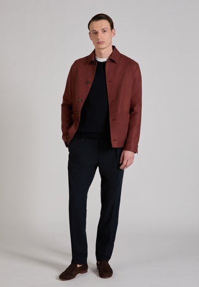 Rust-colored woven jacket with brown buttons, paired with a navy knit sweater and dark trousers. The ensemble features a tailored fit.