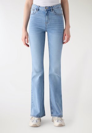 Donna con jeans bootcut a vita alta di colore azzurro chiaro e sneakers bianche, in piedi con le braccia rilassate ai lati, su uno sfondo uniforme.