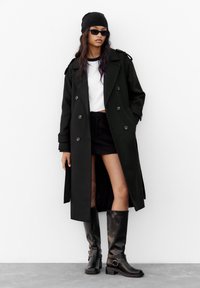 Svart dubbelknäppt trenchcoat med axelremmar, kombinerad med en vit crop top och svart minikjol; glänsande knähöga svarta stövlar.