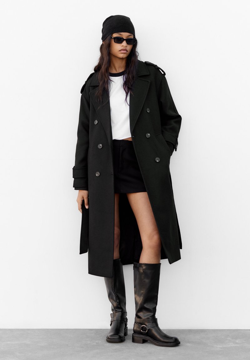 Svart dubbelknäppt trenchcoat med axelremmar, kombinerad med en vit crop top och svart minikjol; glänsande knähöga svarta stövlar.