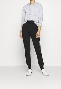 Felpa grigia con tasca frontale e coulisse, abbinata a pantaloni jogger neri con logo laterale e sneaker bianche con dettagli neri.