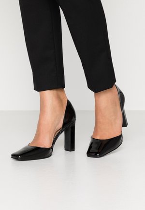 Chaussures à talons hauts en cuir verni noir avec un bout carré et un talon épais. Le design présente un dos ouvert et des accents minimalistes.