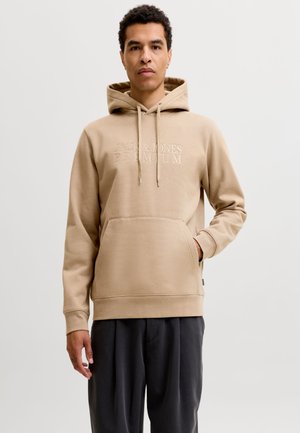 Mann trägt einen beigen Hoodie mit Fronttasche und Kordeln, schwarze Hose, steht mit einer Hand in der Tasche vor einfarbigem Hintergrund.
