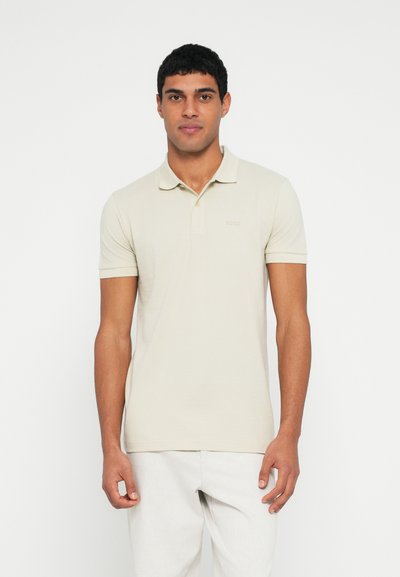 Polo beige à manches courtes en coton, doté d'un col et d'un logo discret sur la poitrine gauche, associé à un pantalon de couleur claire.