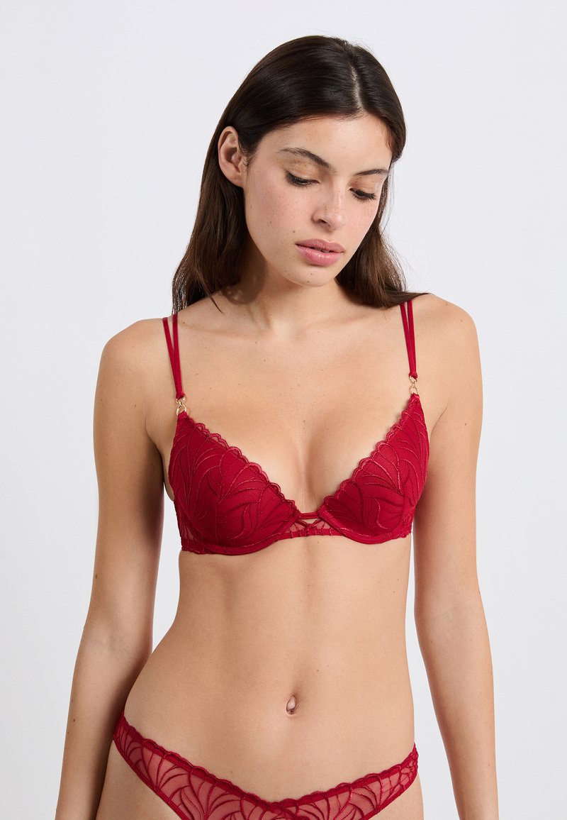 Young woman wearing matching red lace bra and panties with floral patterns, looking down against a plain white background.
