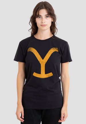 Young woman with brown hair wearing a black t-shirt featuring a large, distressed orange letter Y on the front, standing against a gray background.