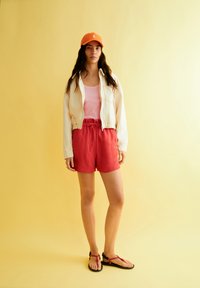 Veste courte beige clair, débardeur rose, shorts rouges avec cordon de serrage, sandales noires avec accents rouges, casquette orange, sur fond jaune.