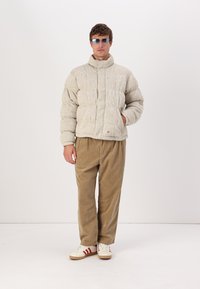 Puffervinterjakke i lys grå struktureret stof, høj krave, quiltet mønster, parret med brune kordfløjspants og hvide sneakers med røde striber.