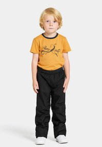 Oranssi puuvillapaita, jossa on musta mustekala -kuvio, yhdistetty mustiin joustohousuihin ja valkoisiin lenkkareihin. Lapsi seisoo valkoista taustaa vasten.