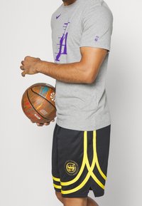 T-shirt Nike gris avec logo violet, associé à un short de basket noir avec des rayures jaunes et un logo, tenant un ballon de basket multicolore.