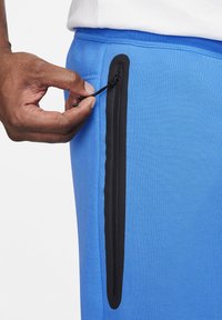 Hand som drar upp den svarta dragkedjan på den vertikala fickan på blå byxor med en vit skjortbakgrund.